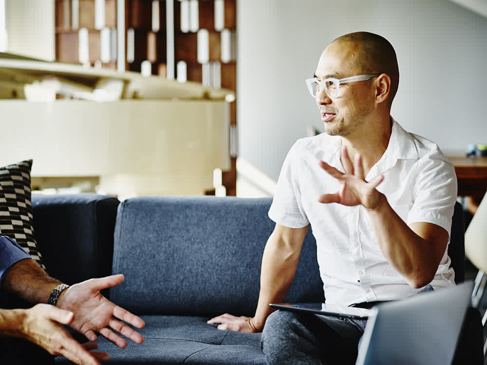 A male employee with shaved hair and white glasses, wearing a short sleeve white button up shirt, sitting on a blue couch expressively talking to another coworker out of frame. 
