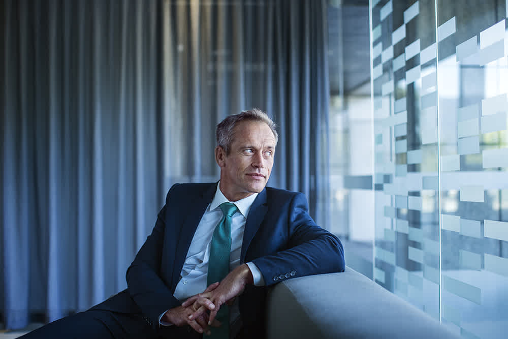 A man in a navy suit with a light blue tie sits on a gray sofa, gazing thoughtfully out of a window. The background features sheer curtains and a textured glass wall.