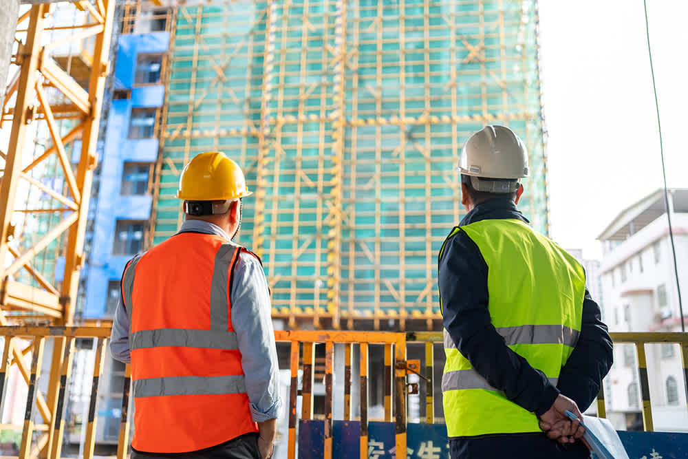 Two construction workers in safety vests and hard hats stand facing a multi-story building under construction. One wears an orange vest, the other a yellow one. Scaffolding and part of a crane are visible in the background, under a clear sky.