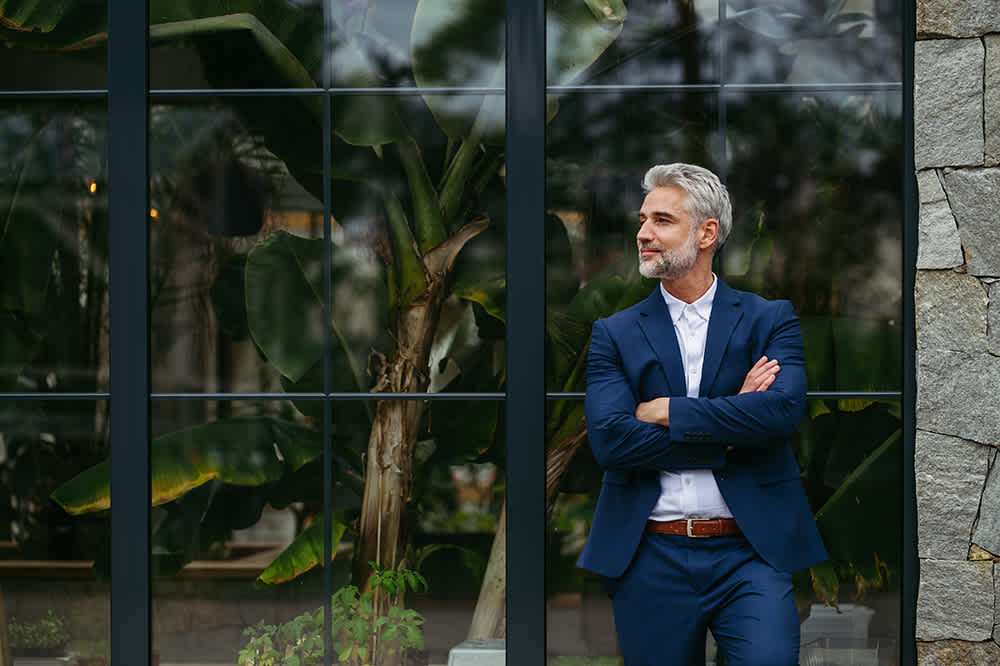 A man with gray hair and a beard stands with arms crossed, wearing a blue suit and white shirt. He is beside a large window with tropical plants visible behind it, next to a stone wall. His expression appears content and thoughtful.