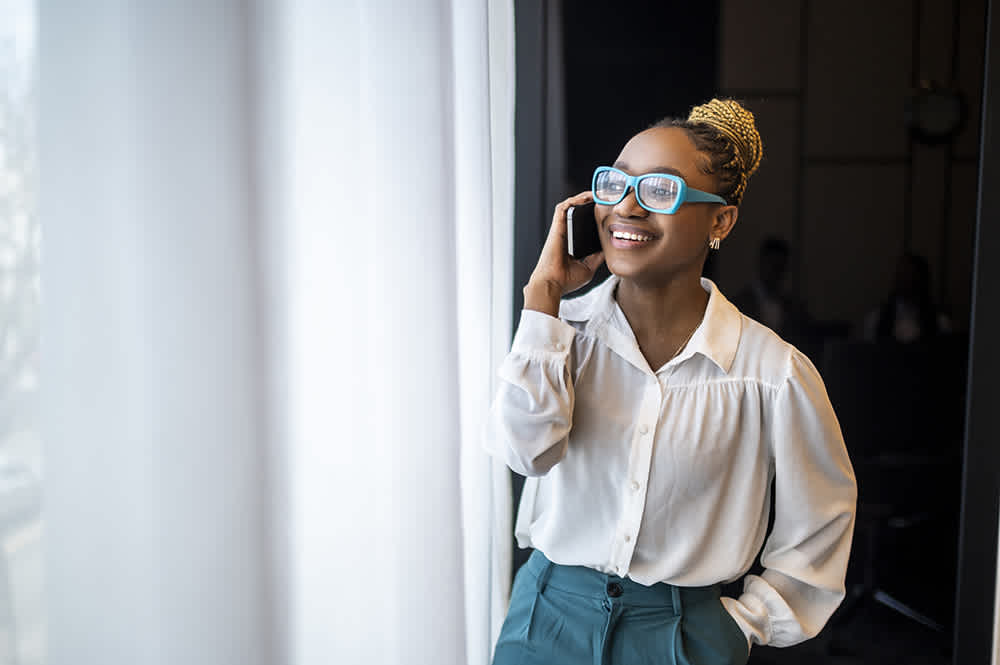 Black woman in teal glasses talking on her phone by a window