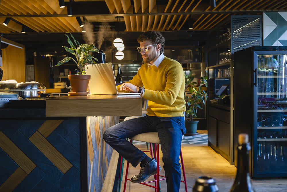 White man restaurant manager with longer, curly hair and glasses wearing a yellow sweater and dark jeans - sitting casually at bar hightop during the day, working on laptop