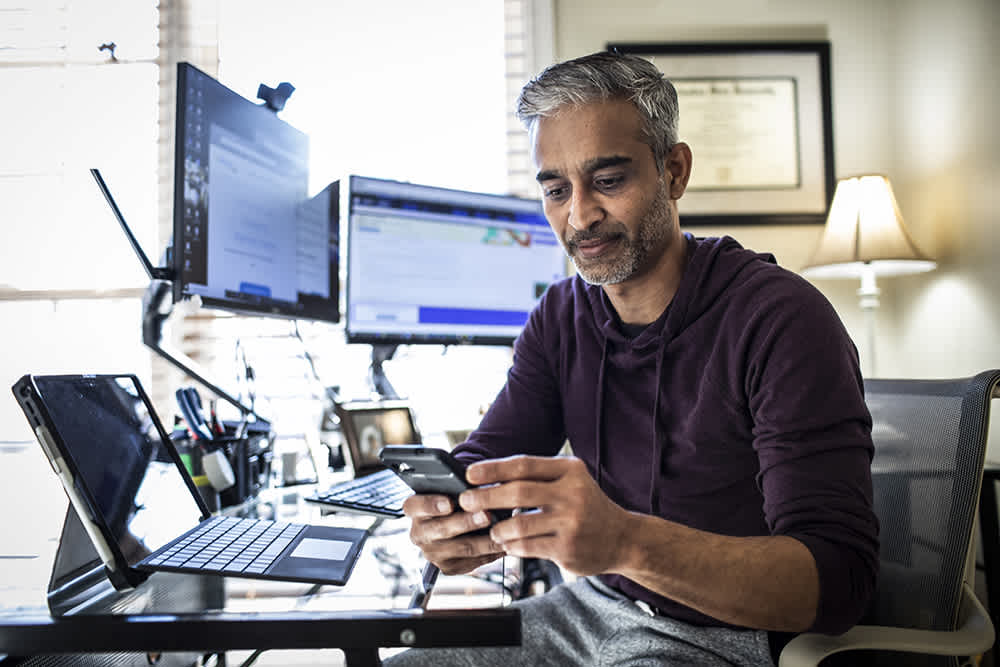Male in front of computers looking down at phone.