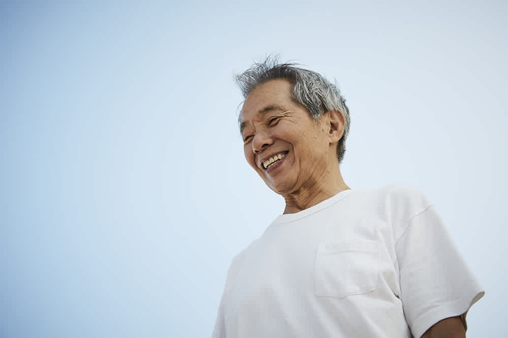 Elderly man in a white t-shirt smiling against a clear blue sky.