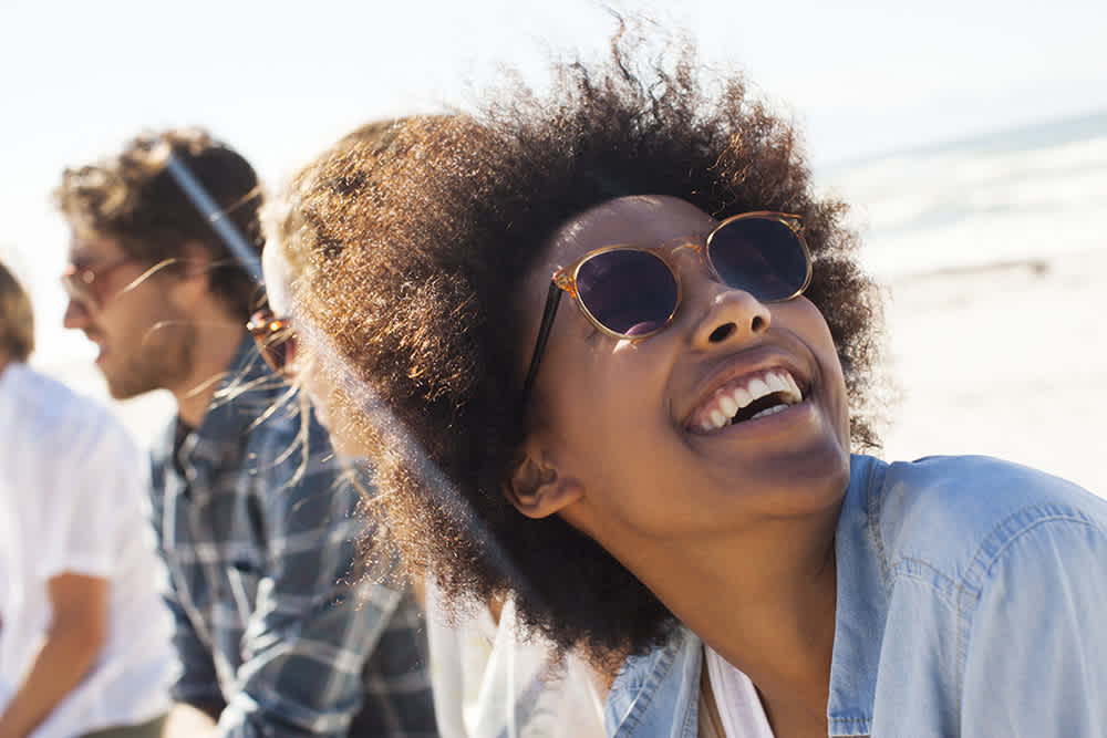 A person wearing sunglasses and smiling looks up while at the beach. They are dressed in a light blue jacket. In the background, another person is partially visible, with a blurred view of sand and ocean.