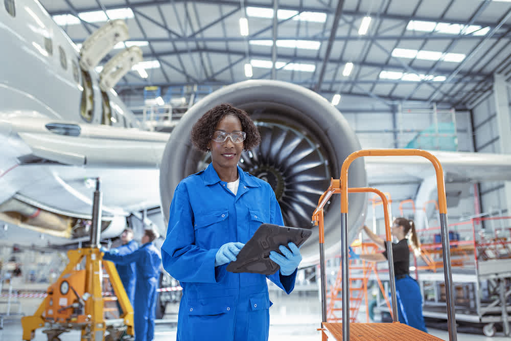 A person in blue coveralls and safety goggles holds a tablet in an aircraft maintenance hangar. They stand in front of a jet engine, with colleagues working in the background. The hangar is spacious and well-lit.