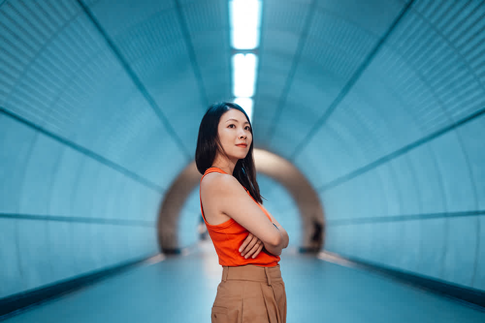 A woman in an orange top and brown pants stands confidently with crossed arms in a modern, blue-lit underground tunnel. The symmetrical lines of the tunnel converge toward a distant point, creating a sense of depth.