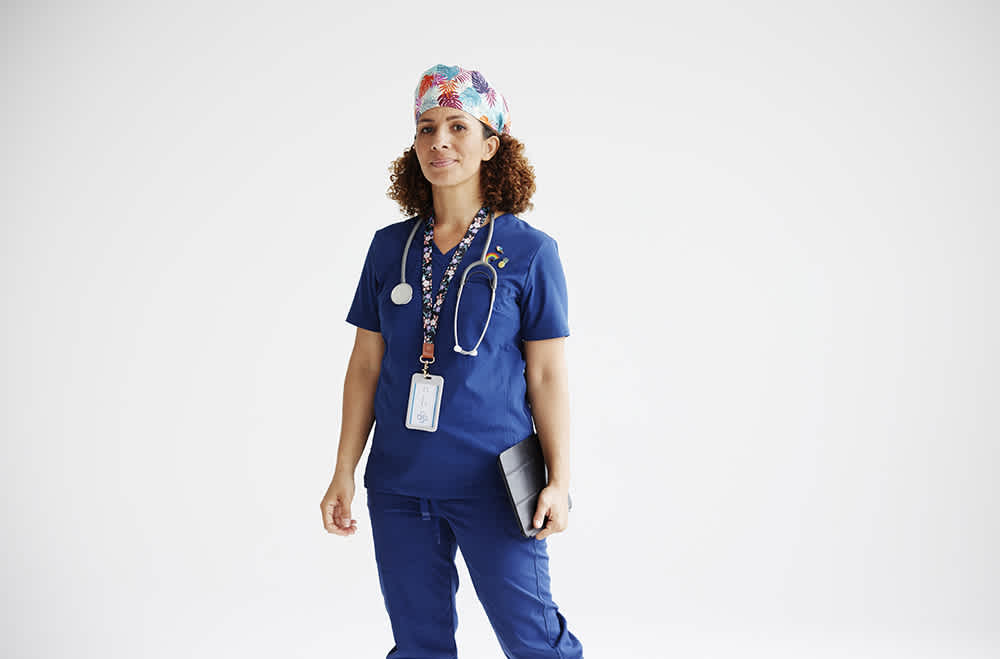 A healthcare professional wearing blue scrubs, a colorful surgical cap, and a stethoscope around the neck stands against a white background. They hold a tablet and wear an ID badge on a lanyard.