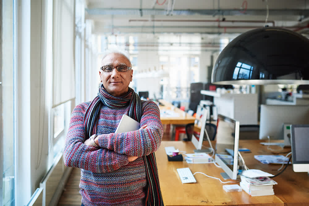 A person with glasses and a colorful sweater stands confidently in a bright, modern office, holding a laptop. The background features large windows, long tables, computers, and a large round black lamp.