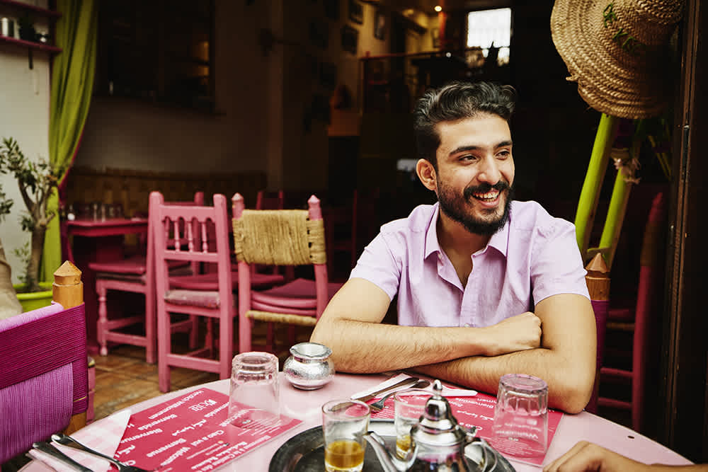 A man with a beard, wearing a light purple shirt, is smiling while seated at a table in a restaurant. The decor features pink chairs and green curtains. Various dishes and glasses are on the table.