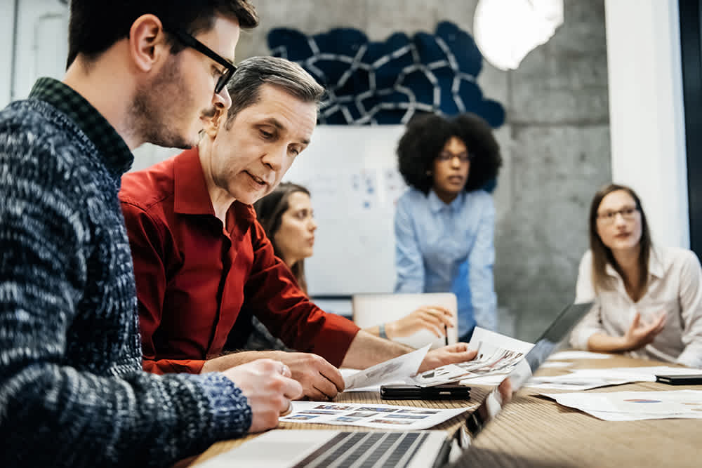 A group of five people sit around a table in a meeting room. They are looking at documents and laptops, engaged in discussion. A whiteboard with a geometric design is visible in the background. The atmosphere appears collaborative.