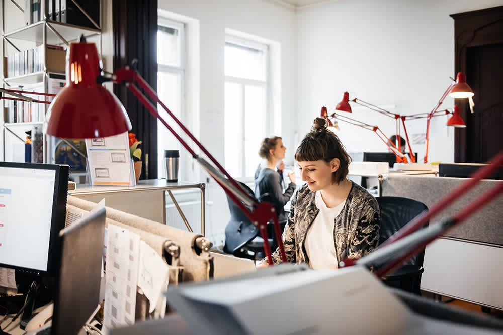 white woman with short brown hair sitting at desk in team office of other desks, computers and red lamps