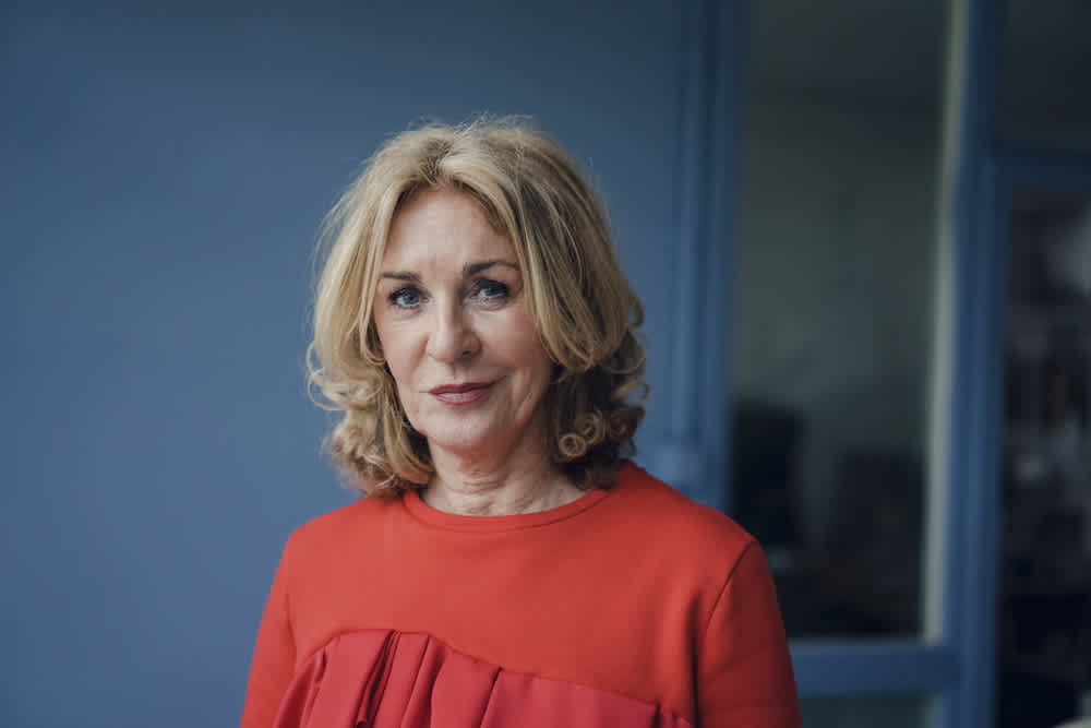 Mid-aged woman with blonde hair and re-orange top standing in front of a navy blue wall.