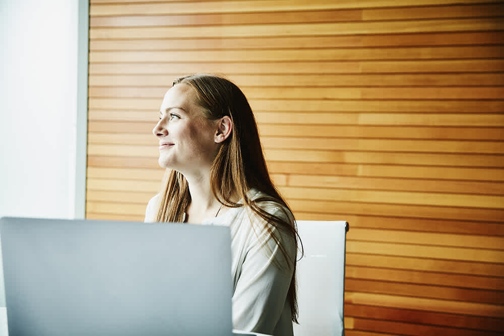 white woman with long hair working on her laptop in front of wooden wall