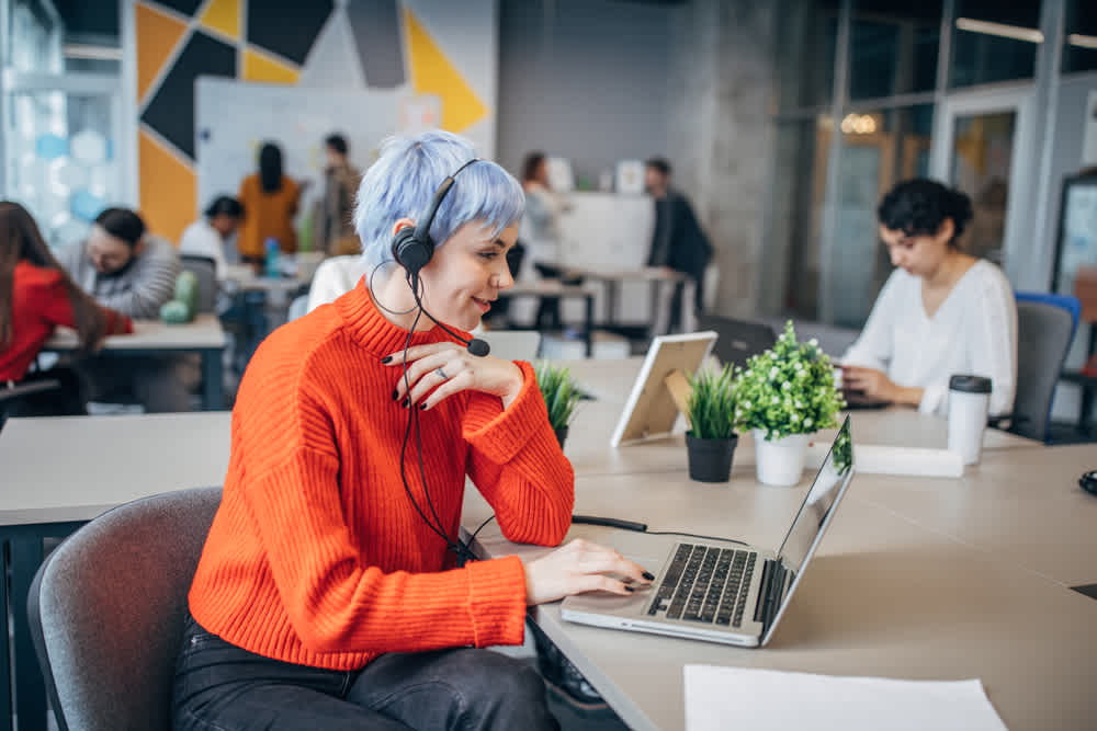 white woman with short white hair in orange sweater, working in an open office 
