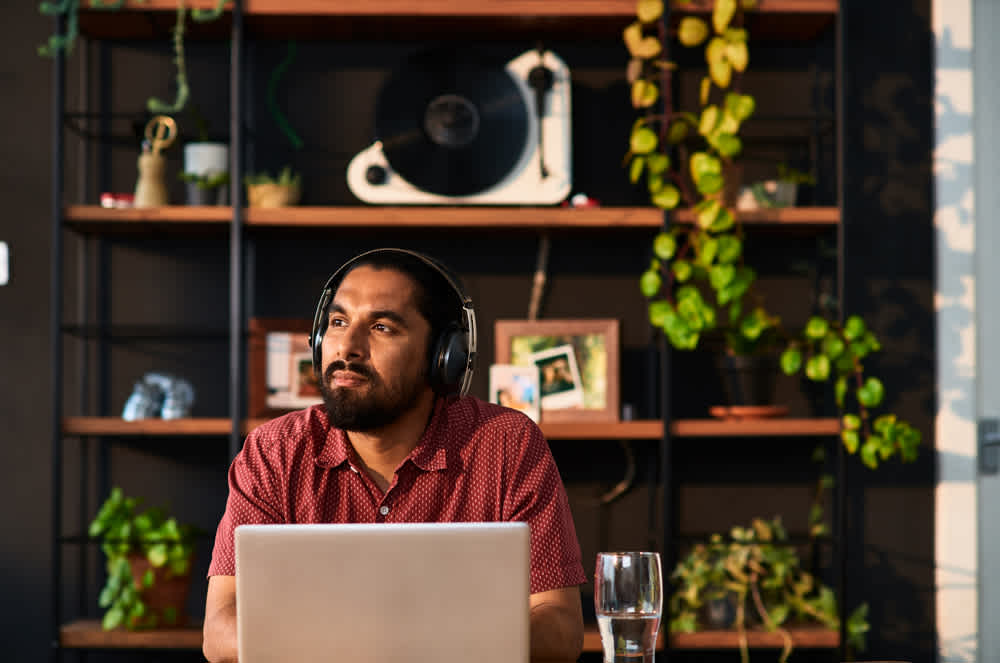A man wearing headphones sits at a table with a laptop in front of him. He appears thoughtful, looking into the distance. Behind him is a shelf with plants, framed photos, and a record player. A glass of water is on the table.
