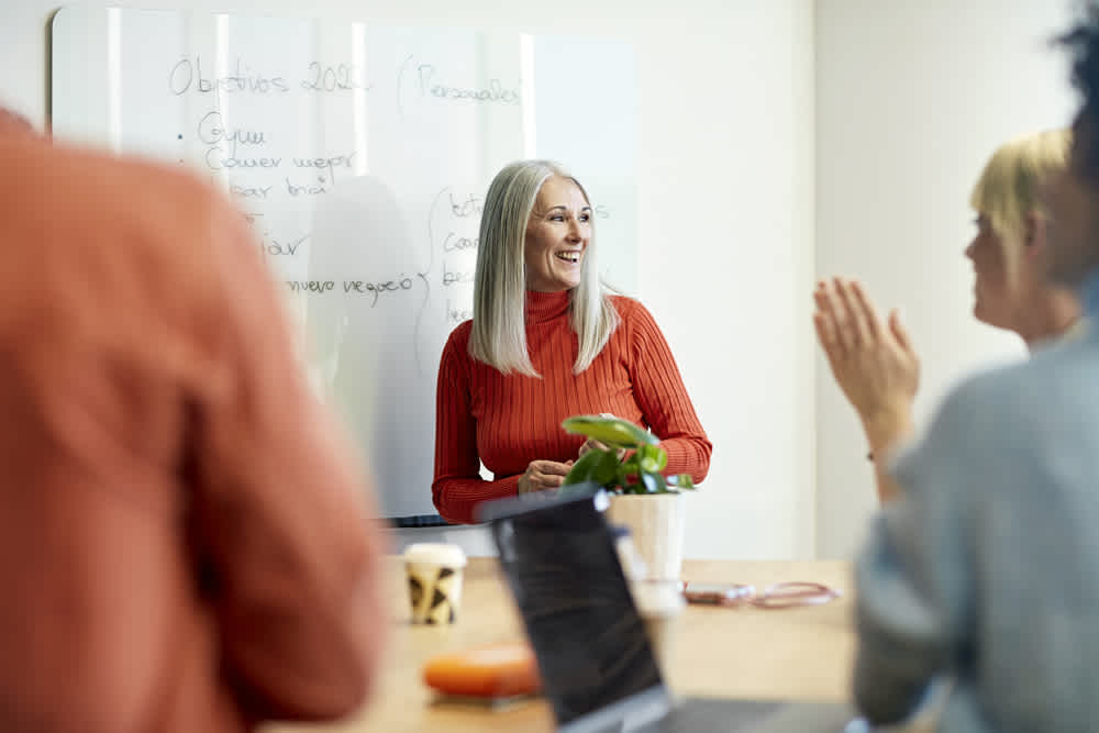 A smiling woman with long gray hair in a red sweater stands in front of a whiteboard, engaging with two people seated and clapping. The table holds a laptop and a plant. The setting appears to be a meeting or presentation.