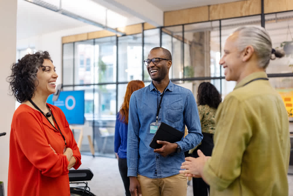 Three people are standing and chatting cheerfully in a modern office with large windows. One holds a tablet. The space has plants and a screen in the background, creating a casual and collaborative atmosphere.