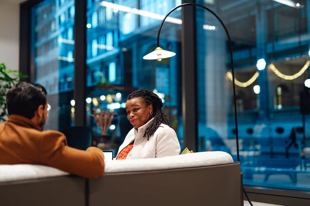 Two people sit on a couch in a modern, well-lit office space with large windows. One person faces away, wearing a brown coat, while the other, wearing a white jacket, smiles and listens. City lights are visible outside.