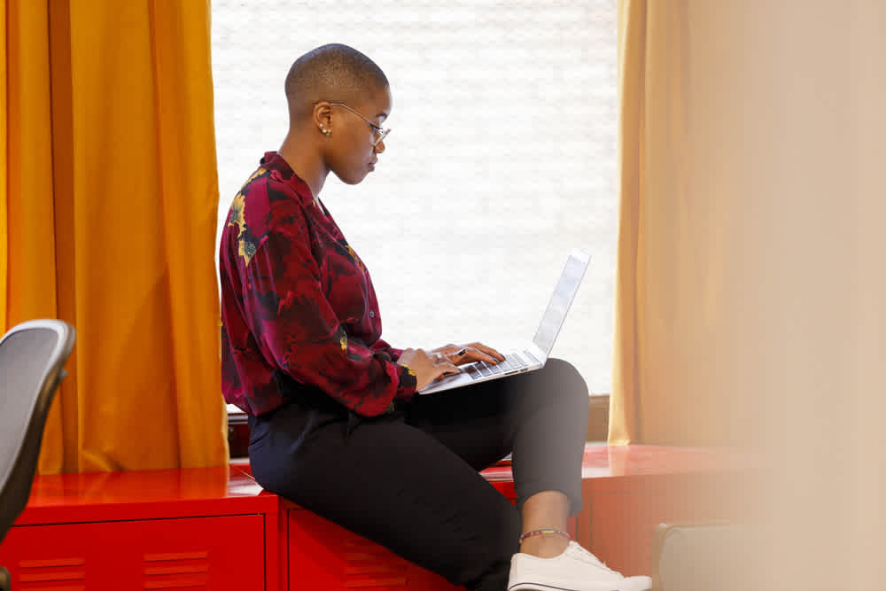 side profile of black woman with shaved hair working on laptop sitting next to window with orange curtains