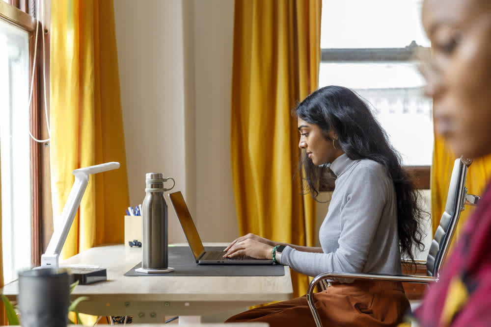 A woman works on a laptop at a desk with a lamp, water bottle, and stationery. She is sitting in a room with yellow curtains and a window, concentrating on her screen. Another blurred person appears in the foreground.