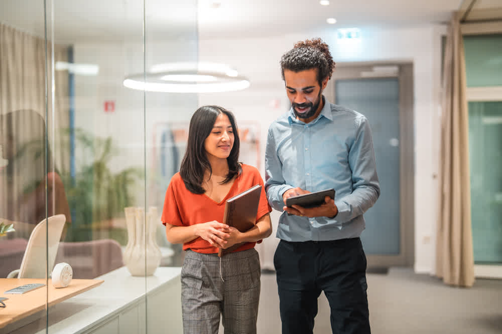 A man and woman walk together in an office hallway. The woman holds a notebook, and the man looks at a tablet. They appear engaged in conversation. The setting is modern, with glass walls and minimalist decor.