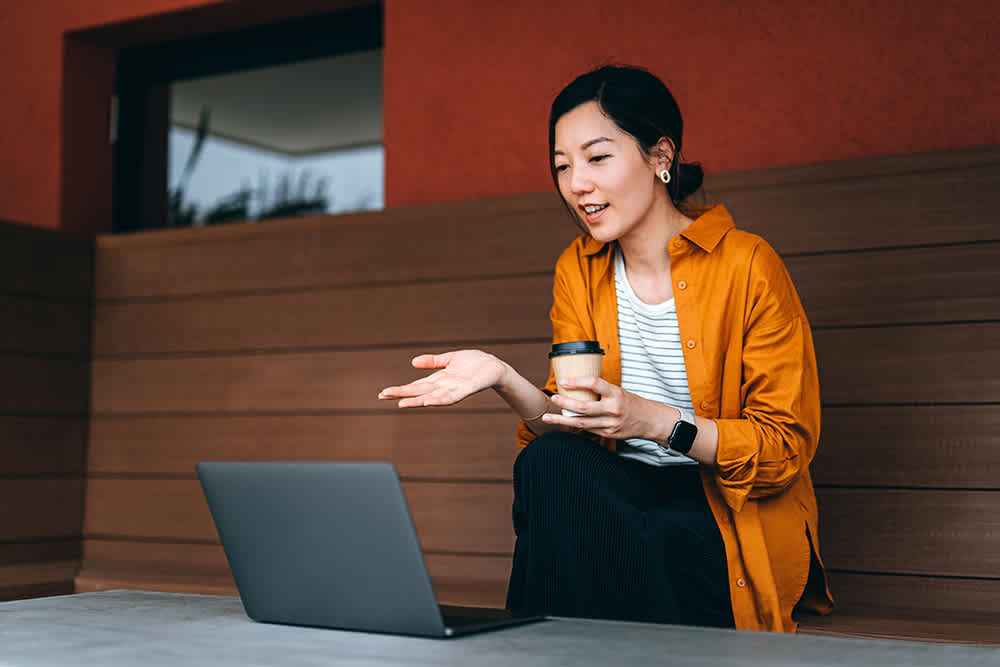 A woman sitting on a wooden bench holds a coffee cup, gesturing towards a laptop on a table. She wears a yellow shirt over a striped top. The setting appears to be an outdoor patio with a red wall in the background.