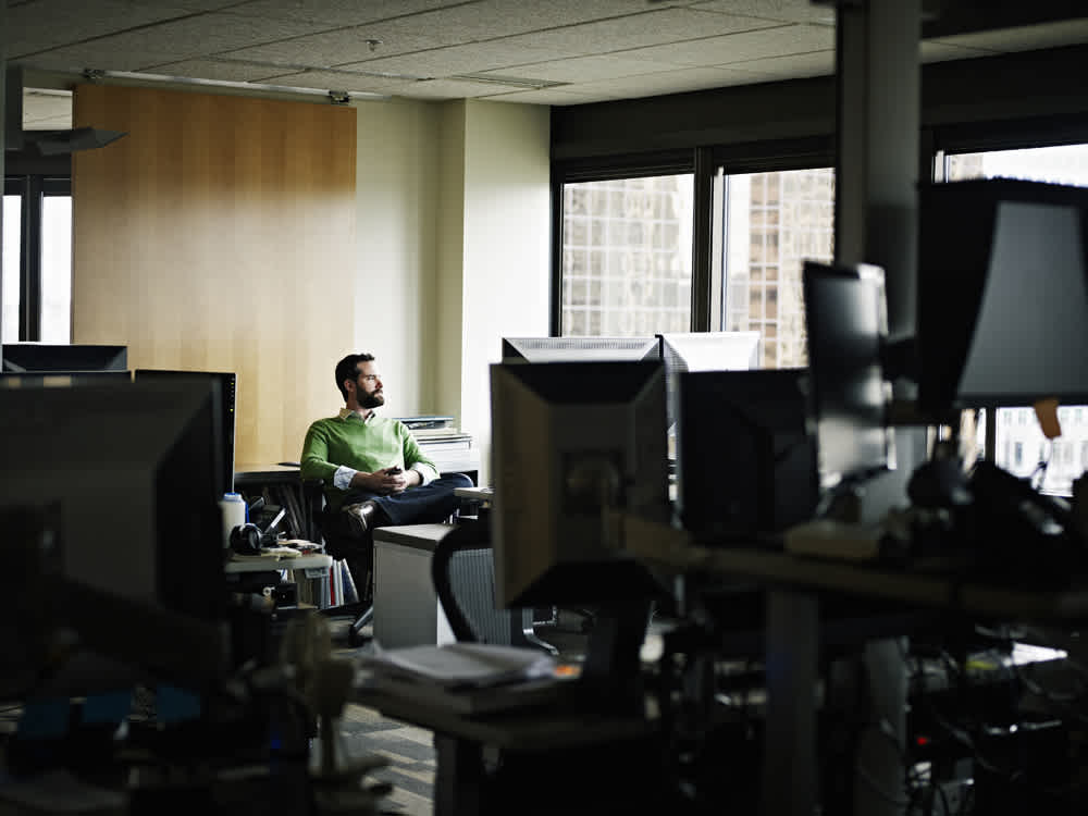 A man in a green sweater sits at a desk in a dimly lit office, looking out the window. The office is filled with computers and office supplies. Sunlight streams through the window, casting a soft glow over the scene.