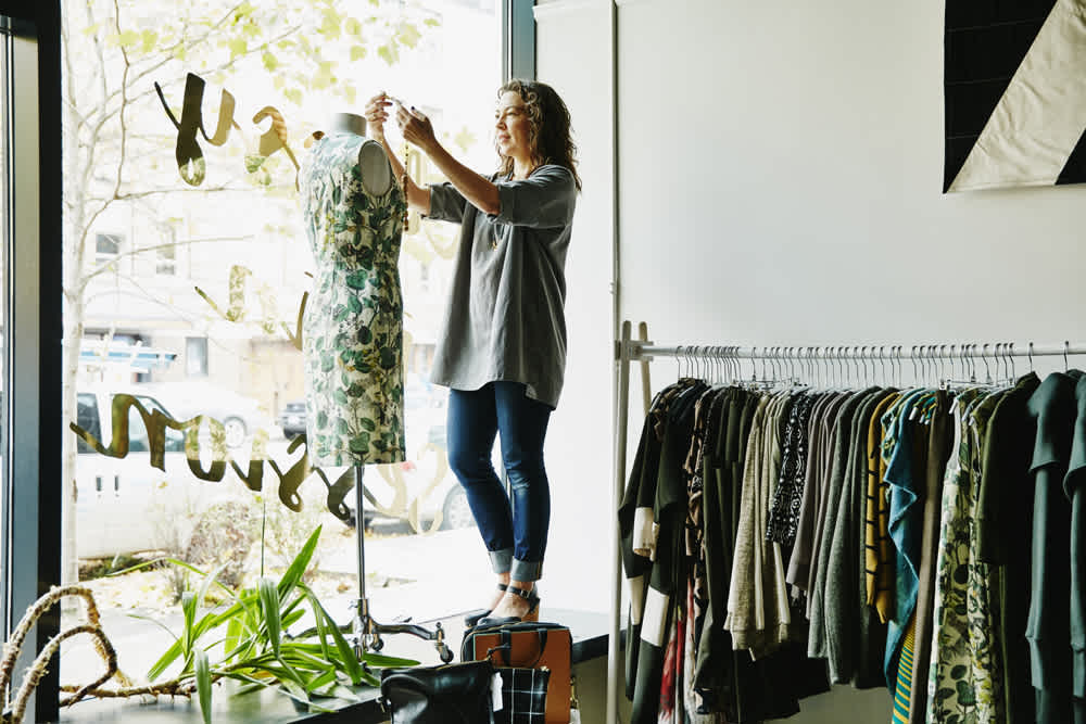 A woman adjusts a patterned dress on a mannequin in a clothing store. She stands on a platform near large windows with city views. Racks of colorful clothing line the wall, and sunlight brightens the modern, stylish interior.