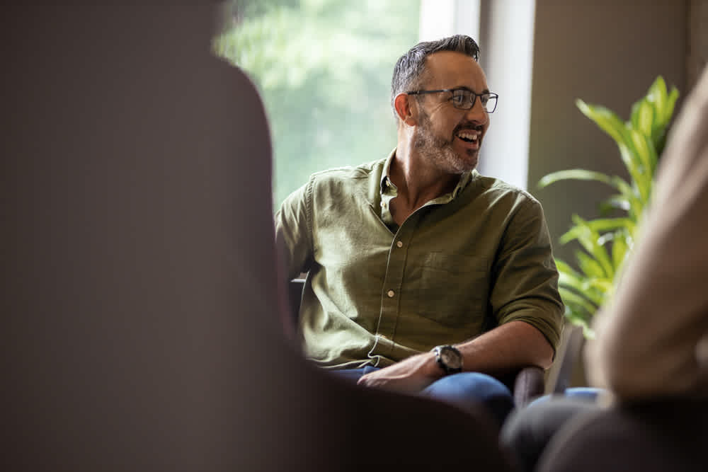 Man with glasses and a beard, wearing an olive green shirt, sits and smiles in a bright room with a large window. He appears engaged in conversation, with a blurred plant in the background.