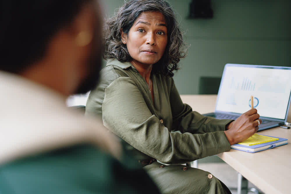 A woman with gray hair sits at a table, holding a pencil and looking at a person across from her. She is in front of a laptop displaying graphs. The setting appears to be a casual office or meeting room.