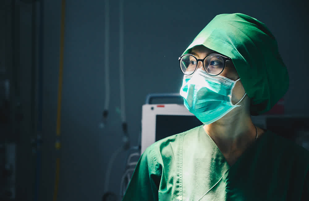 A medical professional wearing a green surgical gown, cap, and face mask stands in a dimly lit room. They are looking to the side, with medical equipment partially visible in the background.