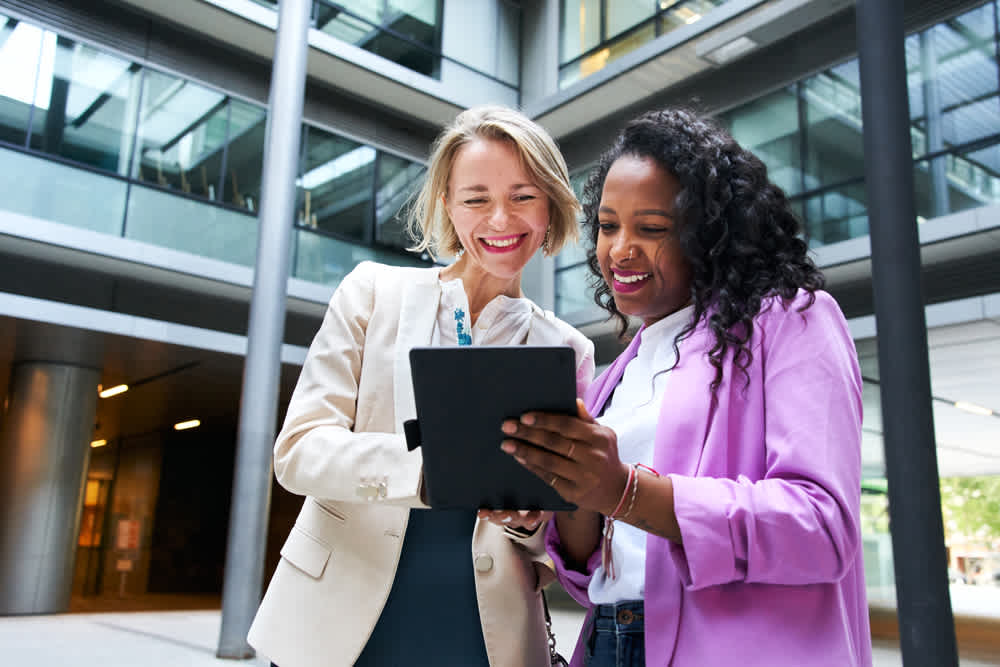 Two women in professional attire smile as they look at a tablet outside a modern office building. One wears a beige blazer, the other a pink one. They appear engaged and happy, with tall glass windows and pillars in the background.