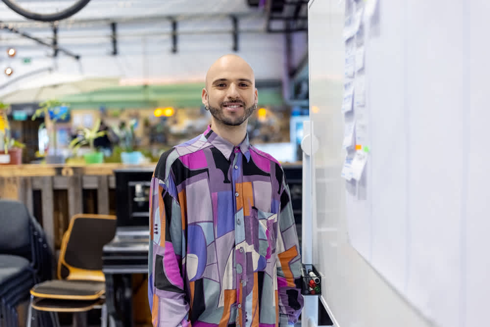 A person with a shaved head and a colorful geometric patterned shirt stands smiling beside a whiteboard covered in sticky notes. The background shows a blurred office space with chairs and plants.