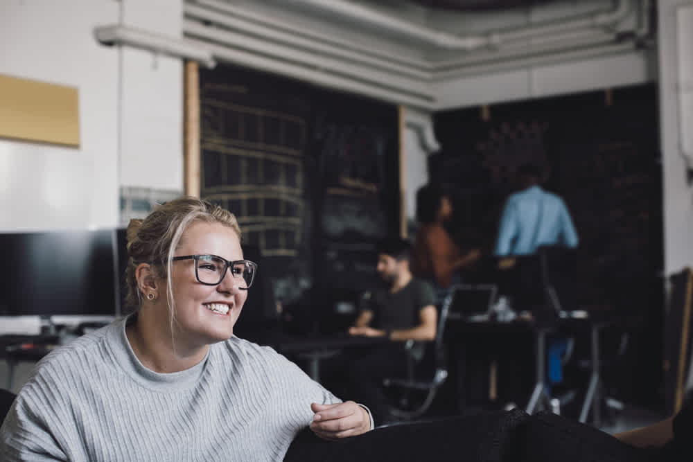 A woman with glasses and a gray sweater smiles, sitting in a dimly lit office with chalkboard walls. In the background, two people stand by a chalkboard while another person sits at a desk with computers.