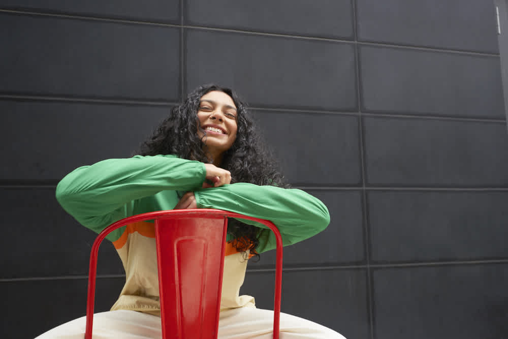 A person with long curly hair smiles while leaning on the back of a red chair. They are wearing a green and beige outfit, seated in front of a dark, textured wall.