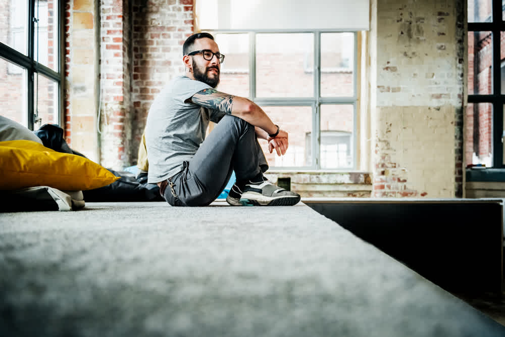 A man with glasses and a tattooed arm sits on a carpeted platform in a modern, industrial-style room with large windows and exposed brick walls. He is wearing a gray t-shirt, dark pants, and sneakers, looking relaxed and thoughtful.