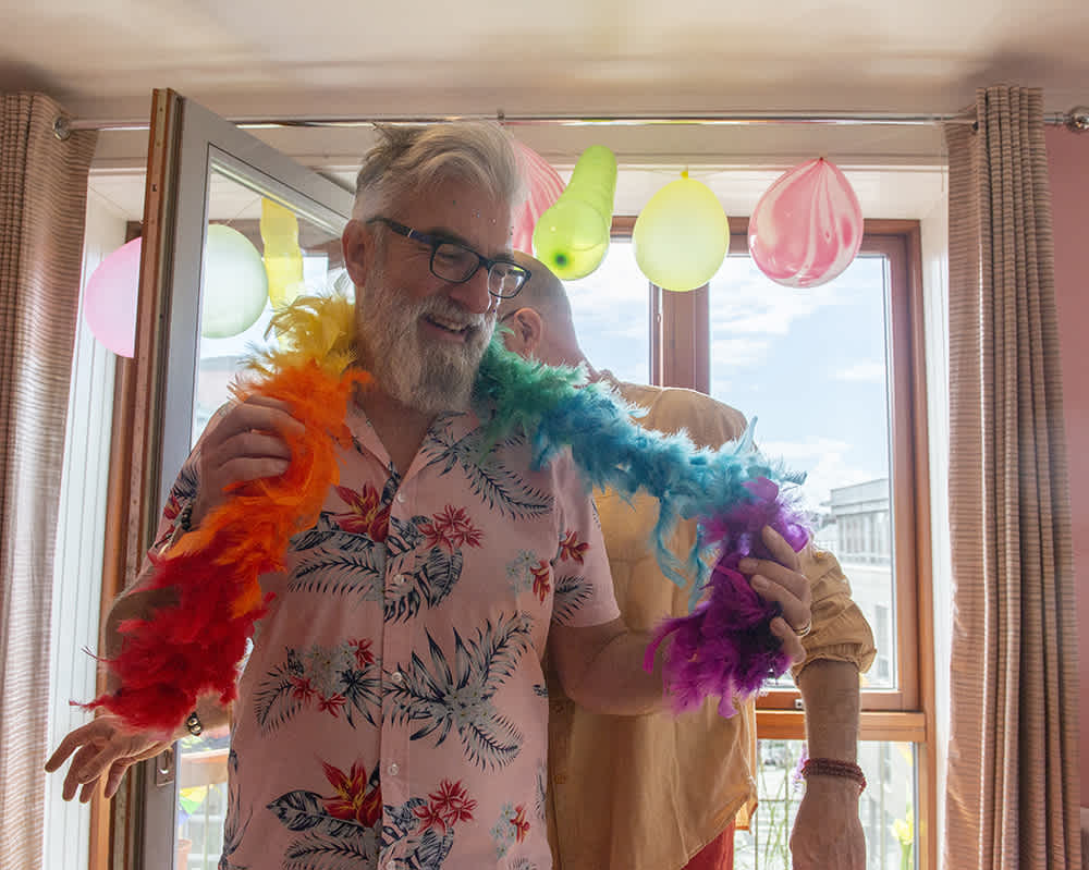 A person with a white beard, wearing a colorful feather boa and a floral shirt, smiles at a party. Balloons are hanging in the background near a window. Another person stands behind them, partially visible.