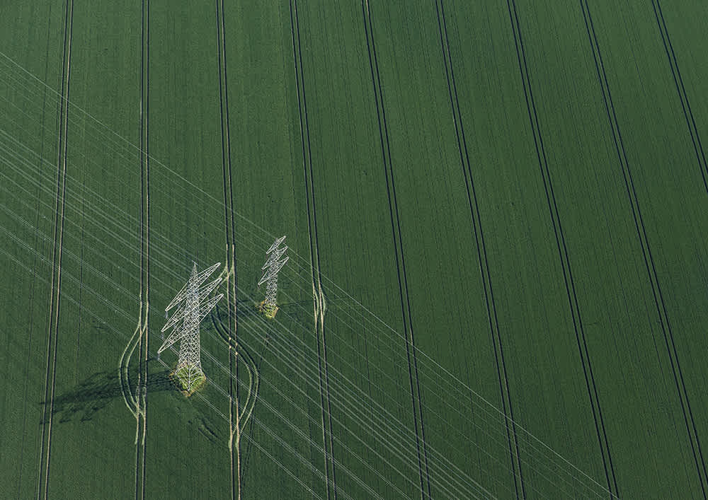 Aerial view of a green agricultural field with intersecting lines of tire tracks. Two transmission towers stand in the field, casting long shadows, with power lines stretching diagonally across the landscape.