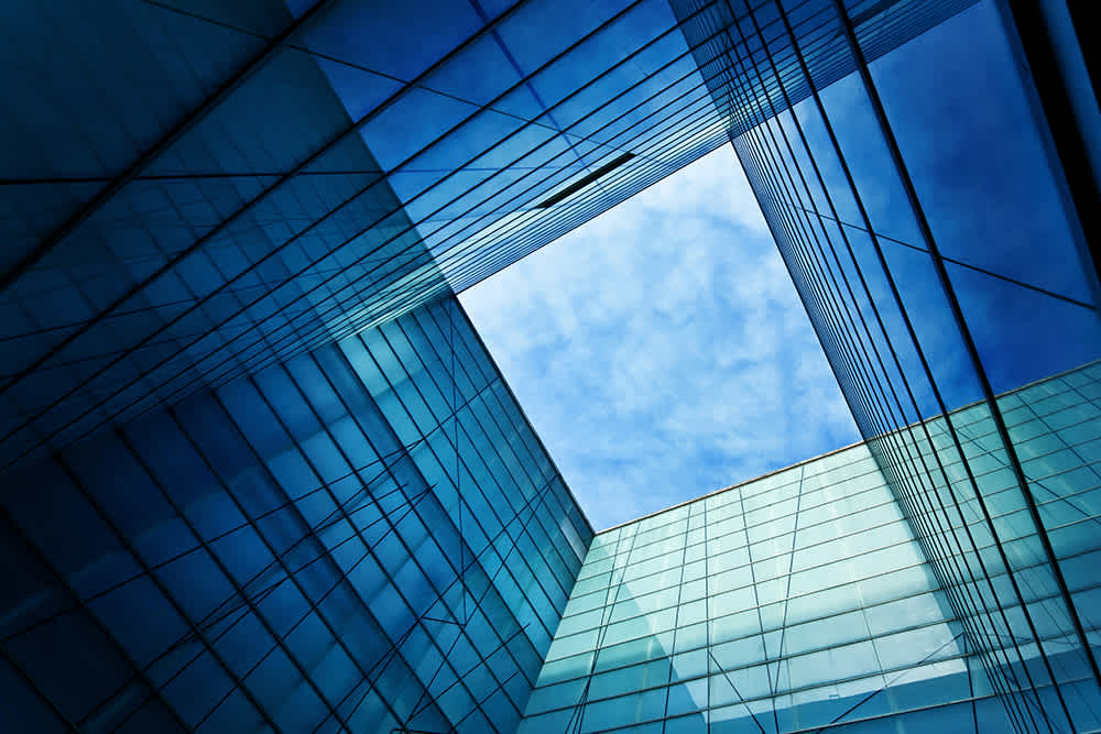 View from below of modern glass buildings forming a square opening to the sky. The reflections on the glass create a geometric pattern, with a clear blue sky visible through the opening.