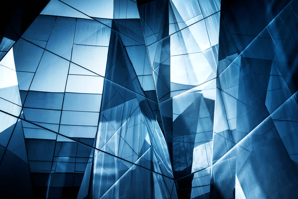 Abstract architectural image featuring a fragmented, geometric glass facade. The structure displays a complex pattern of reflective surfaces and angles, bathed in blue light.