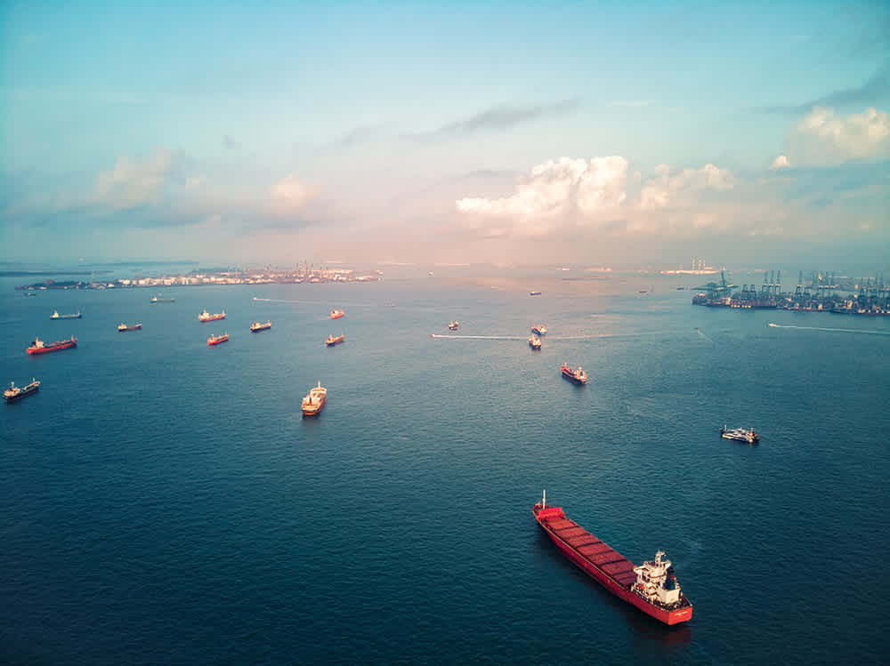 Aerial view of several cargo ships and tankers scattered across a calm blue sea, with a port visible in the background under a partly cloudy sky.
