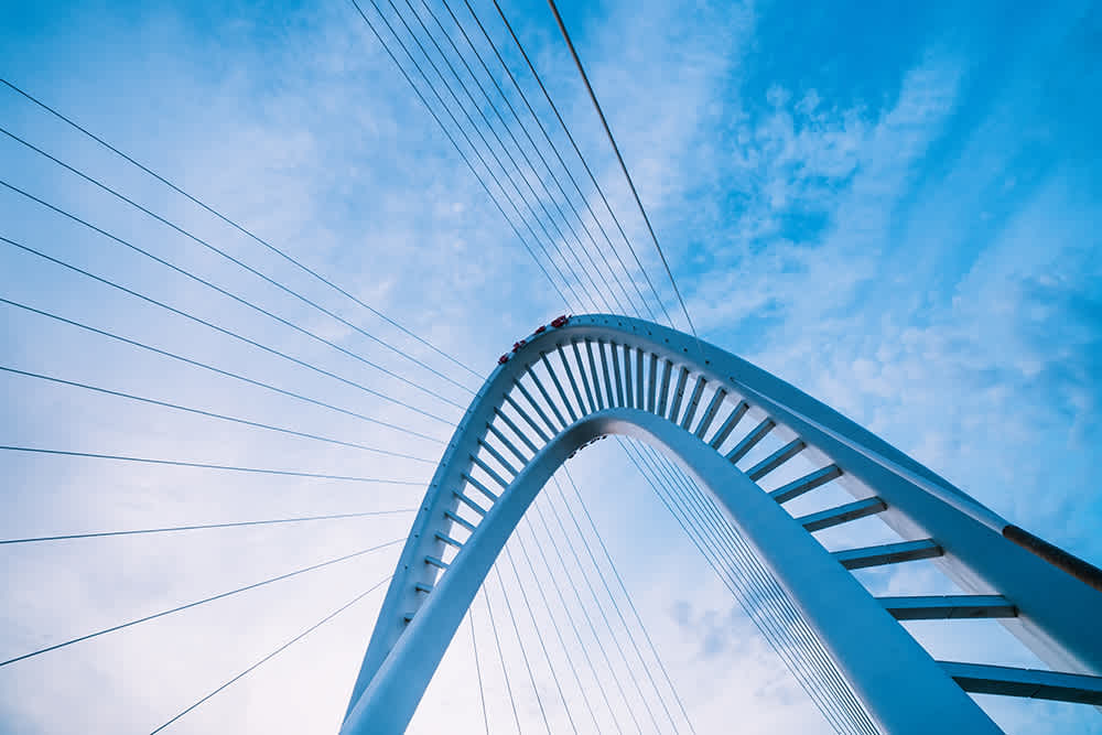 A modern, white suspension bridge with an arched design stretches across the image against a backdrop of a blue sky with light clouds. Thin cables extend from the arch to the bridge deck.