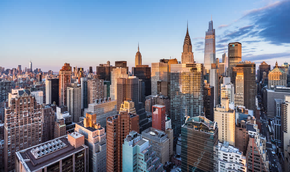 Aerial view of a cityscape at sunset, featuring a dense cluster of tall skyscrapers with modern and classic architectural styles. The sky is partly cloudy with pink and blue hues, and sunlight reflects off the buildings windows.