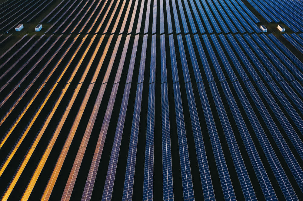 Aerial view of a vast solar farm with neatly arranged solar panels. The panels reflect a gradient of light, creating colorful patterns in shades of blue, orange, and yellow. Small structures are interspersed among the rows.