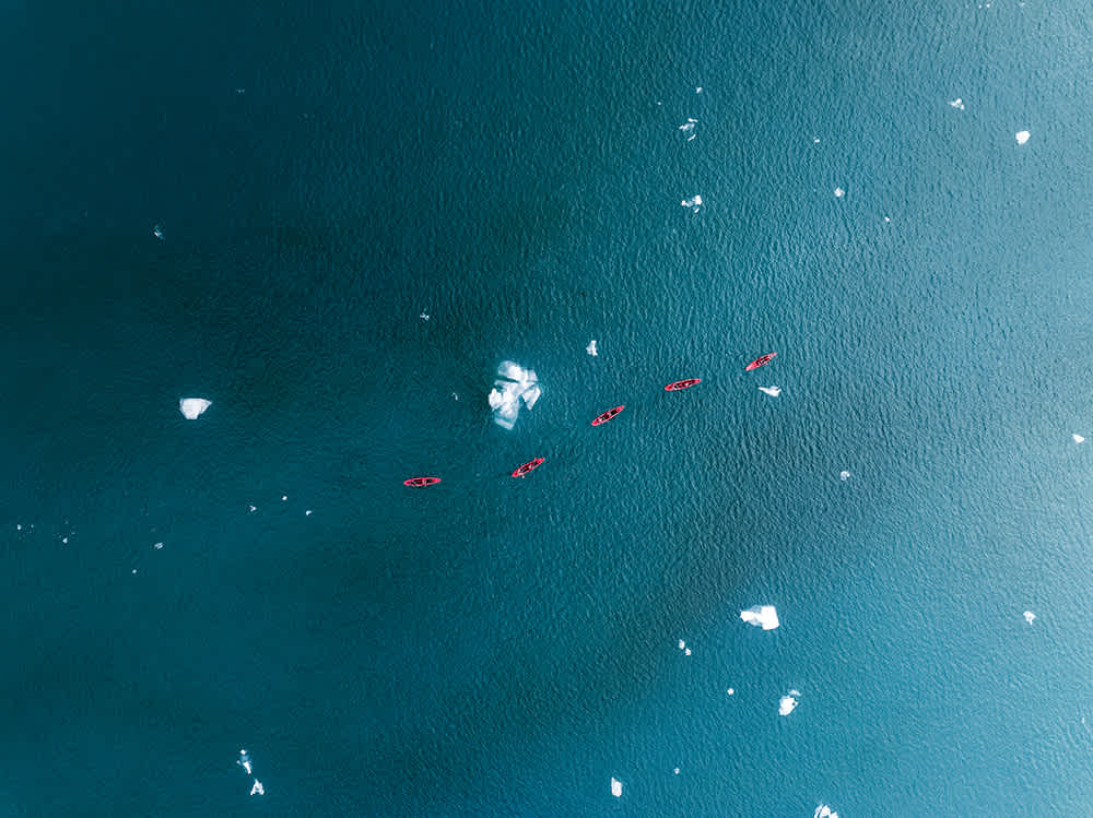 Aerial view of five red kayaks paddling through a vast expanse of teal water, surrounded by small floating ice chunks. The serene setting captures a peaceful exploration of the icy waters.