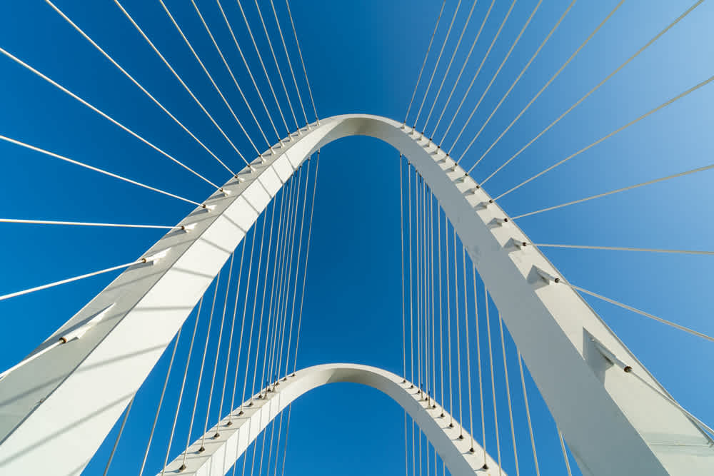 Close-up view of a modern, white suspension bridge against a clear blue sky. Multiple cables fan out from the arch, creating a symmetrical pattern. The perspective emphasizes the height and architectural design of the structure.