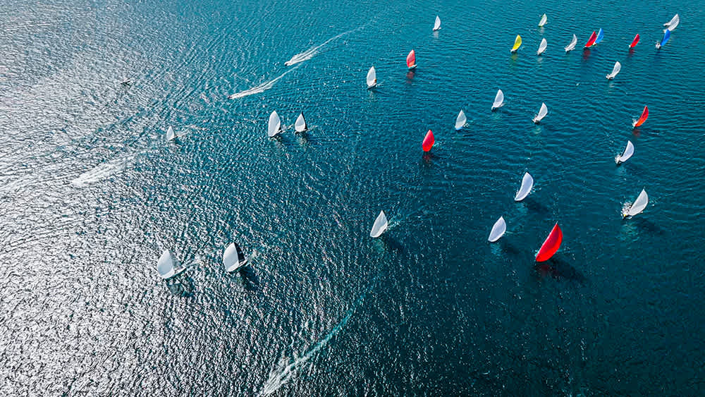 Aerial view of a wide sea with numerous sailboats scattered across the water, some with red and white sails billowing. The sunlight glistens on the rippling waves, creating a sparkling effect.