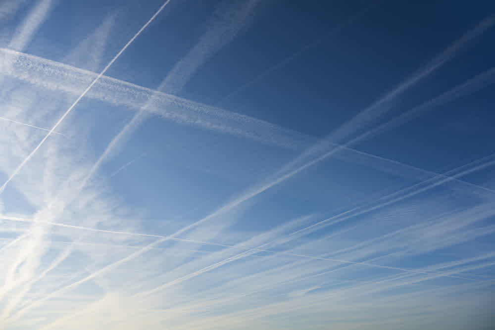 Blue sky filled with numerous intersecting white contrails from airplanes, creating a crisscross pattern. The contrails vary in thickness and blend into the clear sky, with no clouds visible.