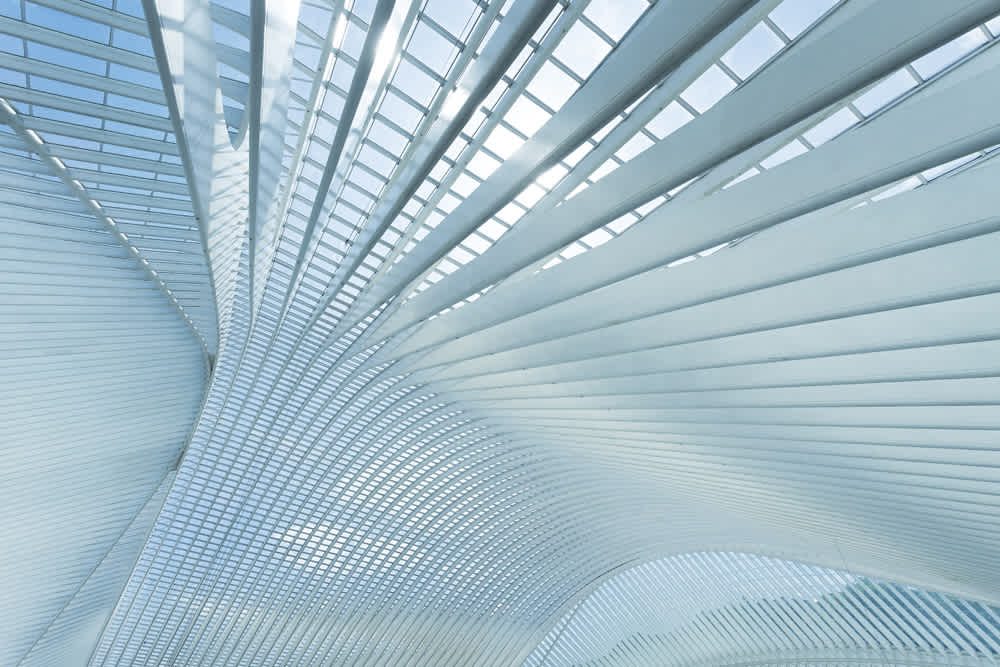 Futuristic architectural interior featuring sweeping, curved metal beams and glass panes. The design creates a dynamic pattern with light streaming through the structure, casting soft shadows and highlighting the airy, expansive space.